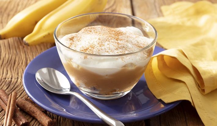Fotografia em tons de branco em uma bancada de madeira de cor marrom. Ao centro, um prato azul contendo um recipiente com a banoffee. Ao lado, há uma colher, um pano amarelo e ao fundo, 2 bananas.