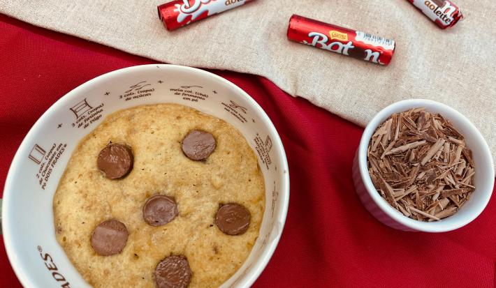 Fotografia em tons de vermelho com um recipiente ao centro. Dentro do recipiente existe um cookie com pedaços de chocolate BATON. Ao lado existe um pequeno pote com raspas de chocolate e ao fundo existe algumas embalagens de chocolate BATON
