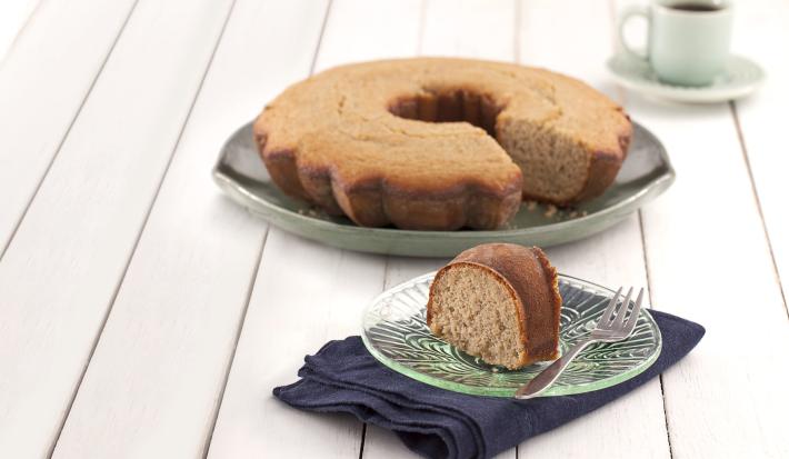 Fotografia em tons de verde em uma bancada de madeira branca, um paninho verde, um prato redondo grande com o bolo de amêndoas e aveia com um pedaço cortado e um pratinho pequeno com a fatia do bolo.