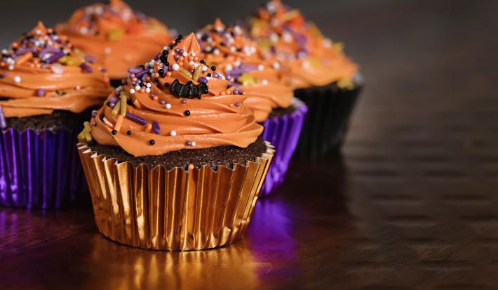 Foto de cupcakes cobertos por um creme laranja, decorados com confeitos coloridos em embalagens brilhantes nos tons de roxo e laranja.