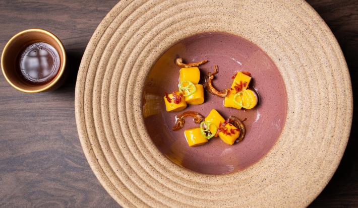 Fotografia vista de cima de um prato grande com o meio fundo. Dentro tem gnocchi de batata doce laranja com dashi de cupuaçu, crocante de jamon e cogumelos. Ao lado do prato tem um recipiente pequeno com molho, que está também sobre uma mesa escura.