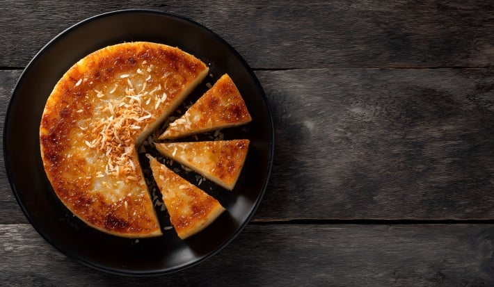 Um bolo de mandioca caramelizado em formato redondo está servido em um prato preto. Algumas fatias já foram cortadas, revelando sua textura cremosa. A cobertura tem um tom dourado e está decorada com coco tostado no centro.