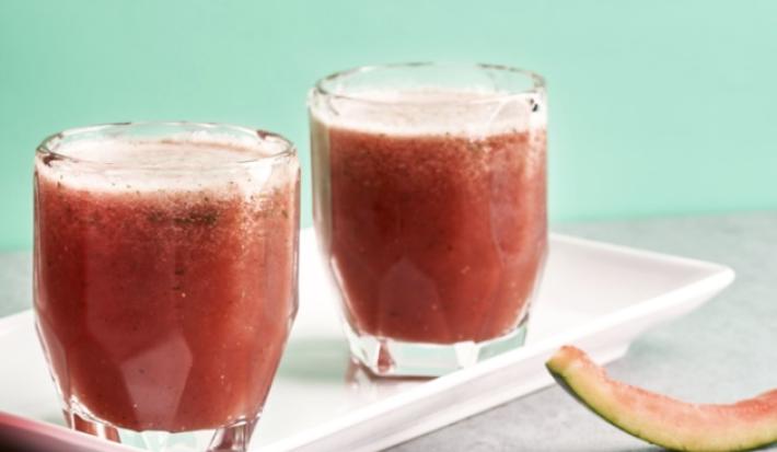 Fotografia em tons de rosa e vermelho em uma bancada de madeira clara com um recipiente branco retangular com o suco de melancia e menta. Ao lado, fatias de melancia e uma melancia inteira.
