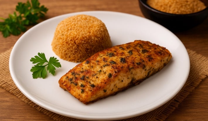Fotografia de um filé de peixe com chibé e três folhas de salsinha em um prato branco e raso. Ao fundo tem mais folhas de salsinha e chibé em um recipiente preto e fundo pequeno.