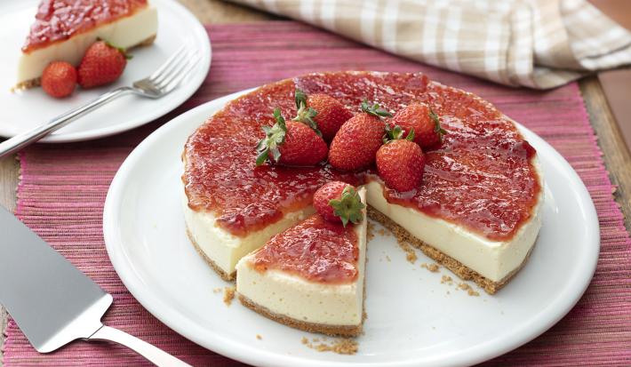 Fotografia em tons de vermelho, em uma bancada de madeira de cor marrom. No centro, uma toalha vermelha e em cima um prato redondo branco contendo o cheesecake. Ao fundo, um pires branco, contendo uma fatia do cheesecake com 2 morangos e um garfo ao lado.