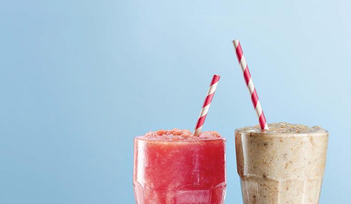 Fotografia em tons de azul e roxo vista de frente. Dois copos transparentes um com uma bebida de cor rosa e o outro com vitamina na cor cinza e ambos contém um canudo vermelho e branco.