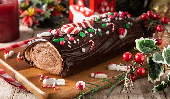 fotografia em tons de marrom, verde e vermelho tirada de uma tábua de madeira. Por cima contém um rocambole de chocolate em formate de troco de arvore com enfeites de Natal.