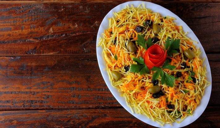 Fotografia de uma salada apresentada em um prato oval branco, com ingredientes finamente picados em tons amarelos e laranjas, azeitonas verdes, batata palha e um enfeite de tomate em forma de rosa, rodeado por folhas de salsa fresca.