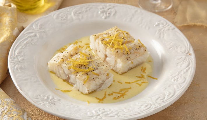 Filés de bacalhau servidos com molho cremoso de ervas e raspas de limão, apresentados em prato branco elegante.