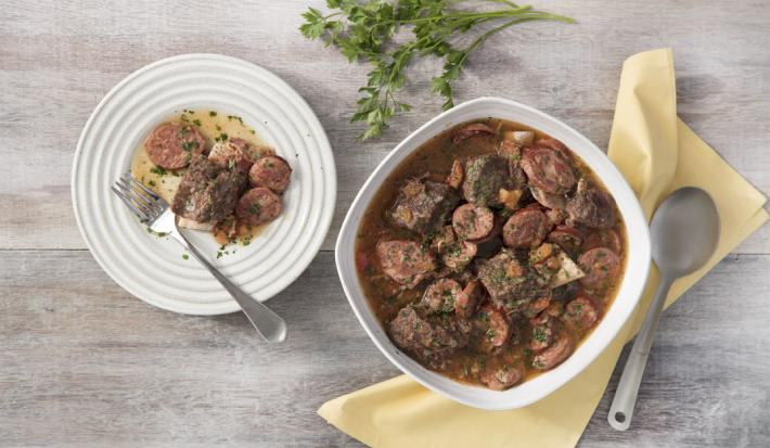fotografia em tons de cinza, branco e marrom de uma bancada cinza vista de cima. Contém um recipiente quadrado branco com pedaços de carne, e ao lado um prato redondo com pedaços de carnes e um garfo para servir.