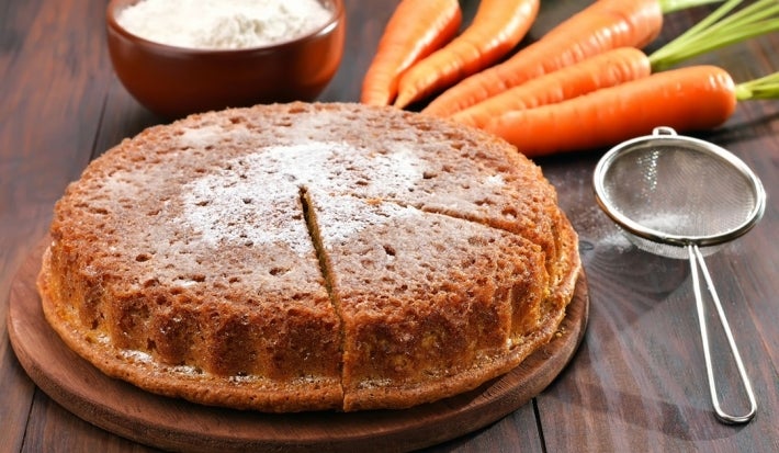 Imagem apresenta um bolo de cenoura em uma tábua de madeira, com uma crosta dourada e uma fatia cortada, revelando seu interior úmido. Cenouras frescas e uma tigela de farinha estão ao fundo, destacando a natureza caseira e apetitosa do bolo.