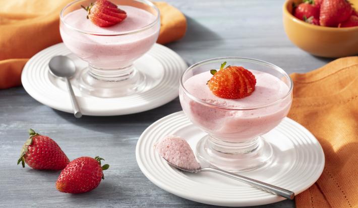 Imagem aproximada da receita de Mousse de Morango Zero Lactose, em tom rosa claro, servida em potes de vidro, decoradas com morangos fatiados. A receita está sobre pratos brancos em uma bancada com um tecido laranja e mais morangos inteiros decorando