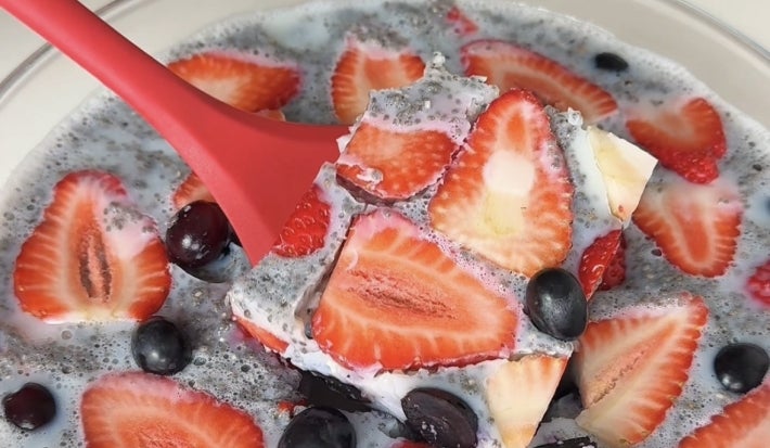Fotografia em tons claros de uma espátula vermelha segurando uma porção da gelatina de frutas e chia, que está dentro de um recipiente de vidro.