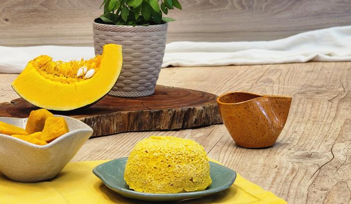 Fotografia em tons de amarelo com um prato azulado ao centro. Em cima do prato existe um pão de cor amarelada feito com abóbora e farinha de aveia. Ao fundo existe uma fatia de abóbora.