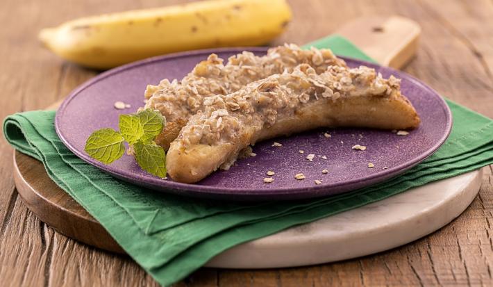 Foto da receita de banana grelhada recheada servida em um prato roxo sobre um paninho verde em uma tábua de madeira de forma circular. Ao fundo há uma banana para decoração