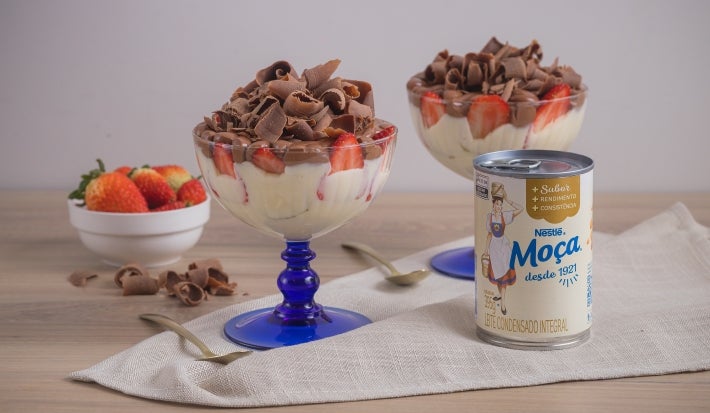 Foto da receita de pavê de leite moça com morangos e chocolate, servidos em taças com uma base azul, sobre uma bancada de madeira decorada com um tecido, pedaços de chocolate, um pote com morangos e uma lata de Leite Moça