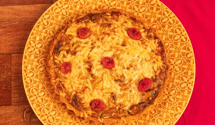 Fotografia mostra uma quiche com queijo gratinado e pedaços de tomate sobre um prato amarelo. Ao fundo, uma mesa de madeira e um pano vermelho.