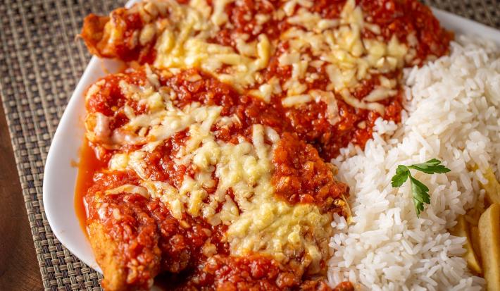 Fotografia de um prato retangular branco com dois filés de frango à parmegiana com queijo derretido por cima, ao lado de um pouco de arroz e batatas fritas.