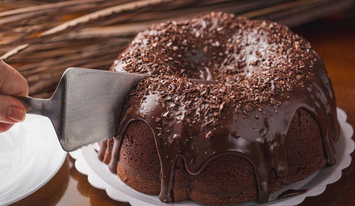 Imagem de um bolo redondo de chocolate com bastante cobertura de brigadeiro e uma mão com uma espátula cortando o bolo.