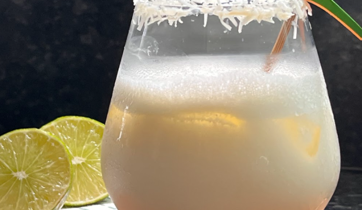 Sobre uma bancada de madeira, um copo transparente com um líquido branco dentro e um pouco de espuma. O copo está decorado com as bordas de coco ralado e um mexedor em formato de papagaio. Ao lado do copo, duas rodelas de limão.