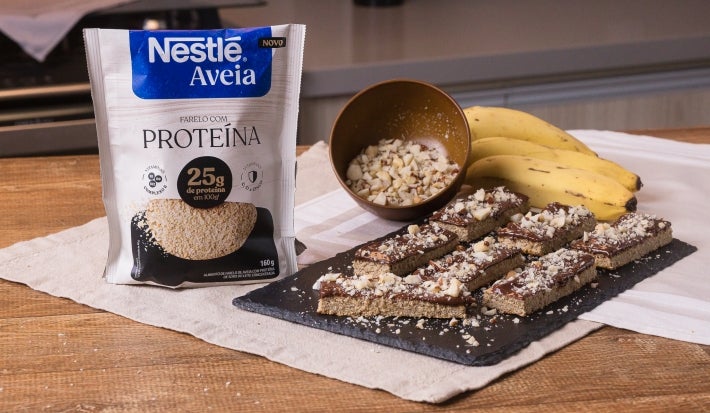 Foto da receita de barrinha proteica de aveia servida em diversas porções sobre uma pedra preta em cima de uma mesa de madeira com um cacho de banana ao fundo