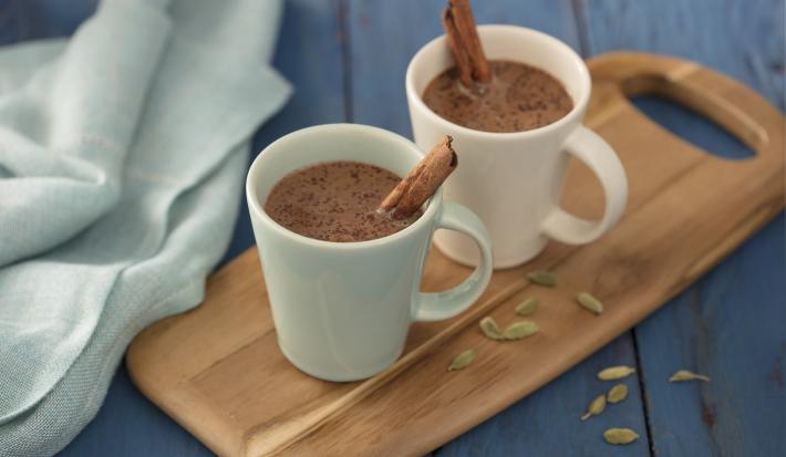 Fotografia em tons de azul em uma bancada de madeira azul, um pano azul, uma tábua de madeira com duas xícaras brancas com o chocolate quente com canela dentro delas.