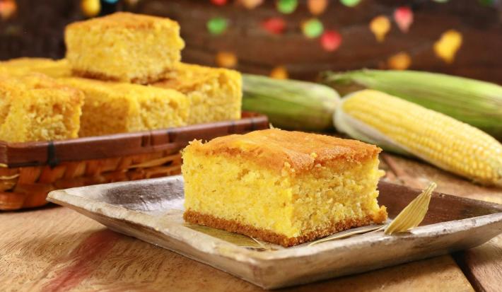 Fotografia de um apoio quadrado de madeira raso com um pedaço de bolo de milho. No fundo tem mais pedaços dentro uma cestinha de madeira, alguns milhos inteiros ao lado e uma decoração de festa junina.