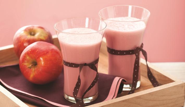 Fotografia em tons de rosa e vermelho, ao centro bandeja de madeira contendo dois copos com creme rosa e decorados com fitas marrom, duas maçãs, sobre guardanapo marrom com borda rosa, tudo sobre bancada de madeira em pátina e fundo rosa.