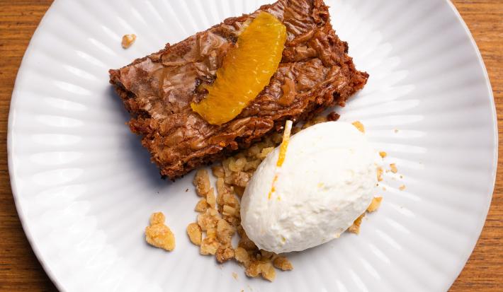 Fotografia de um prato branco raso com um pedaço de bolo de chocolate cremoso com crocante de nozes, e por cima, chantilly de laranja. O prato está sobre uma bancada de madeira clara.