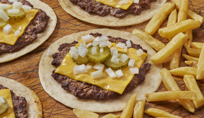 Fotografia mostra uma porção de tacos abertos, sob uma mesa de madeira, com recheio de carne bovina, queijo, cebola, alface e picles para finalizar.