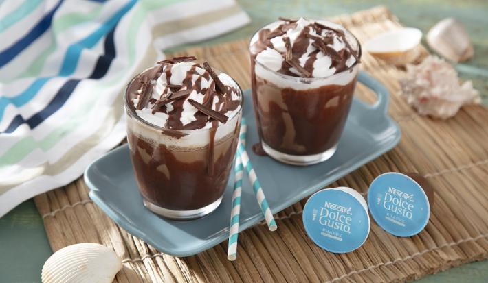 Foto da receita de frapê mocha servida em duas xícaras de vidro em cima de uma base de porcelana azul clara sobre uma esteira de madeira com cápsulas de dolce gusto frapê ao lado