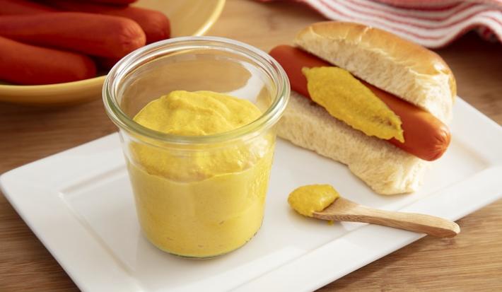 Foto da receita de mostarda caseira com maggi. Observa-se um potinho transparente com a mostarda e, ao lado, um hot dog com o molho passado na salsicha. Também há uma colherinha de madeira com um pouco do molho.