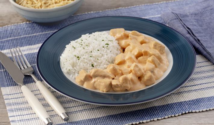 Fotografia em tons de azul em uma bancada de madeira com um jogo americano azul com listras brancas, um prato fundo azul escuro com o strogonoff de frango e arroz branco. Ao fundo, um potinho com batata palha.