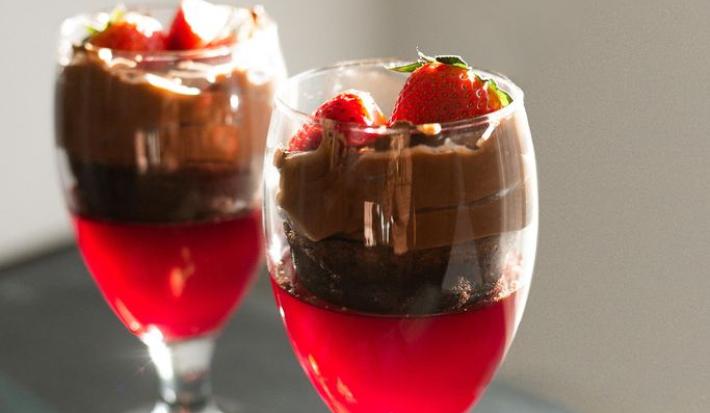 Fotografia em tons de vermelho em uma bancada de madeira cinza escura com duas taças de vidro com o doce de morango com uma camada de brownie de chocolate, outra com mousse de chocolate e finalizando com morangos para decorar.