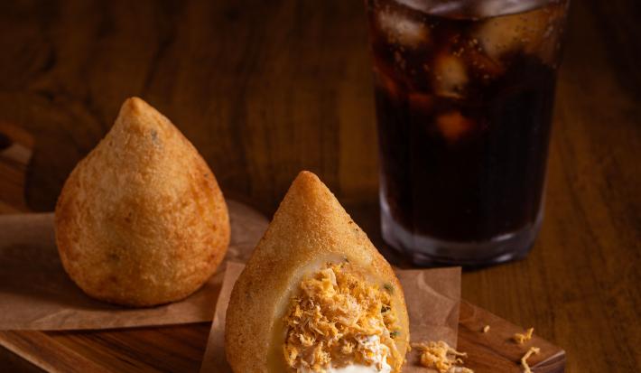 Fotografia em tons de marrom com uma tábua de madeira ao centro. Em cima da tábua existe uma receita de coxinha recheada com frango e requeijao