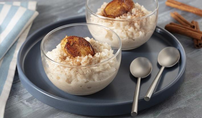 Fotografia em tons de cinza, em uma bancada de madeira escura com um pano azul listrado. Ao centro, um prato redondo preto, com duas taças de arroz doce com banana caramelizada e ao fundo, algumas canelas em pau espalhadas.