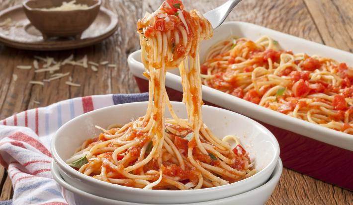 Fotografia em tons de vermelho em uma bancada de madeira com um pano branco com listras vermelhas e azuis, um prato fundo branco com o macarrão ao molho sugo sendo puxado por um garfo. Ao fundo, um recipiente com macarrão e um potinho de queijo ralado.
