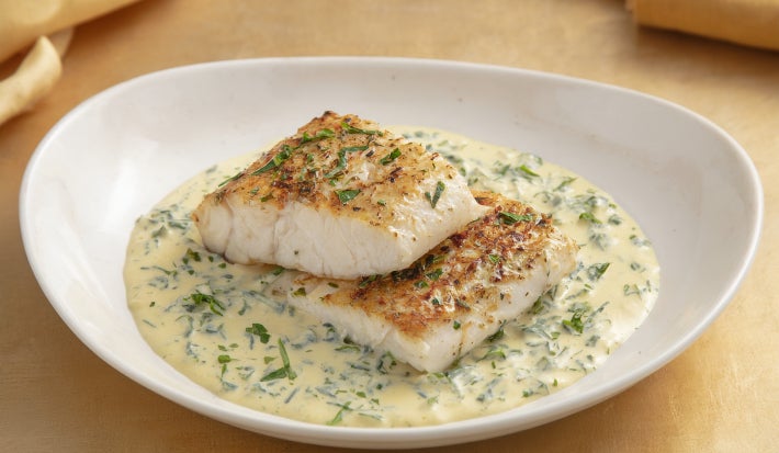Filé de peixe branco dourado servido no molho cremoso de ervas, apresentado em prato fundo branco com acabamento sofisticado.