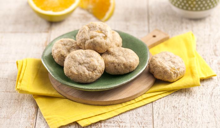 Fotografia em tons de amarelo em uma bancada de madeira clara, um pano amarelo, uma tábua de madeira com um prato verde redondo com alguns biscoitinhos de banana com laranja em cima dele. Ao fundo, uma laranja cortada ao meio e uma xícara verde.