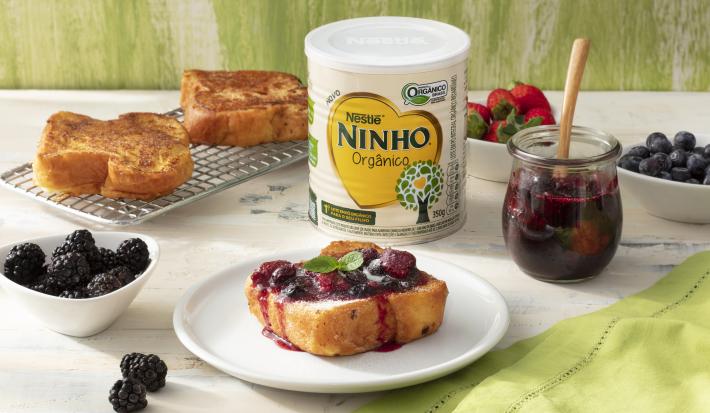 Fotografia em tons de branco e verde de uma bancada branca com um prato branco, sobre ela uma rabanada com geleia. Ao lado um recipientes com frutas vermelhas, uma gradinha com rabanadas, um pote de geleia, uma lata de NINHO orgânico e um paninho verde.