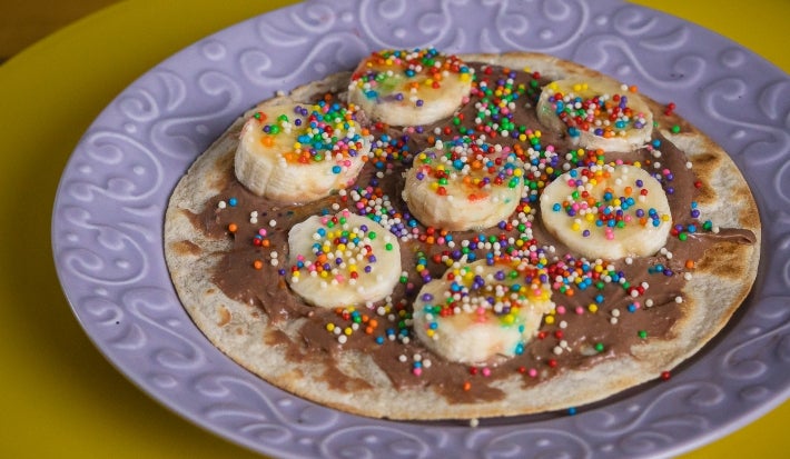 Fotografia de um prato roxo com a mini pizza com chocolate com rodelas de banan e granulado colorido.