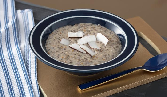 Foto em tons de marrom da receita de mingau de sementes e coco servida em um prato fundo preto com uma colher prateada ao lado sobre uma tábua de madeira lisa com um paninho azul ao lado