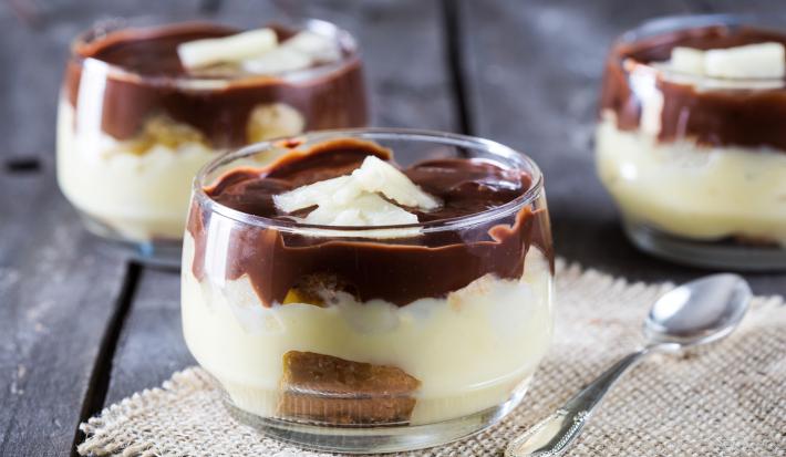 Fotografia em tons de chocolate e branco, com três potinhos contendo um creme branco com biscoitos e coberto com chocolate, um deles sobre toalhinha branca com uma colher ao lado, tudo sobre bancada de madeira em tom roxo.