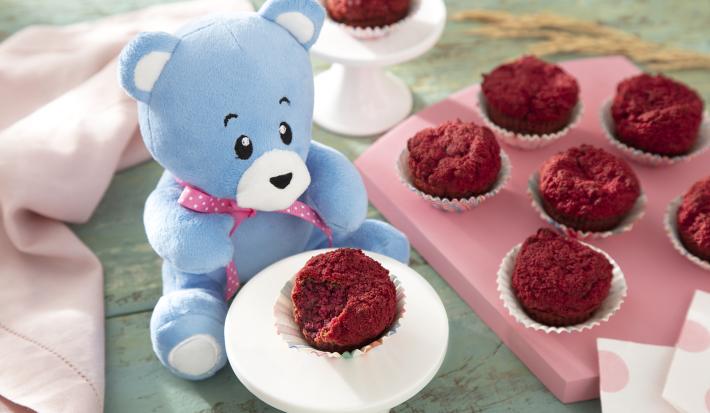 Foto da receita de Muffin de Beterraba. Observa-se uma tábua rosa com 6 muffins e, ao lado esquerdo, um pratinho branco com outro muffin rosa. O ursinho Bo está atrás desse pratinho branco.