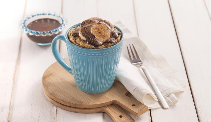 Foto da receita de bolo de caneca com cacau e banana em uma caneca azul, sobre tábuas de madeira, com um guardanapo de pano branco e um garfo sobre ele.