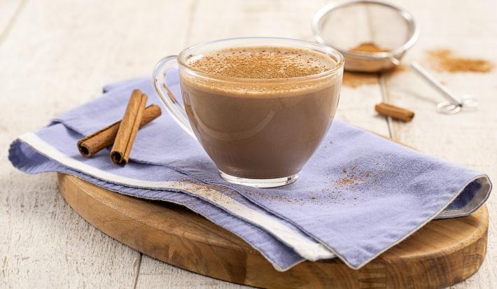Foto da Receita de Cappuccino Cremoso. Observa-se uma xícara de vidro transparente sobre um guardanapo azul e uma tábua de madeira. Ao fundo, uma peneira com canela em pó.