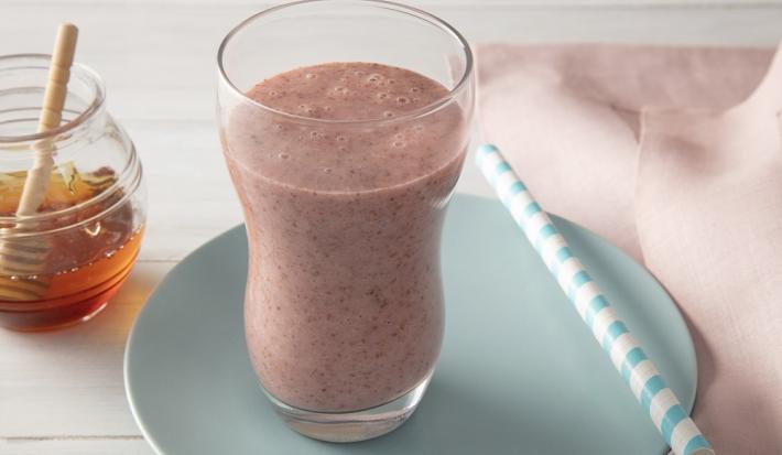 Foto da receita de smoothie iogurte molico com morango e chia servida em um copo largo sobre um pratinho azul com um canudo listrado ao lado junto deum paninho rosa. Ao fundo há um vidro de mel