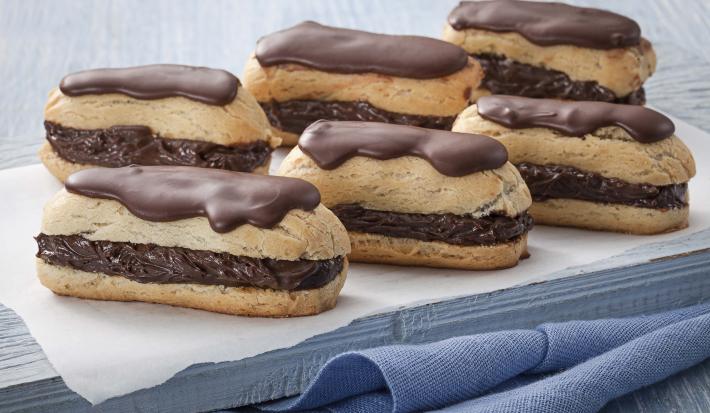 Fotografia em tons de azul em uma mesa de madeira azul com um paninho azul e por cima um guardanapo branco de papel e seis bombas de chocolate com cobertura de chocolate em cima.