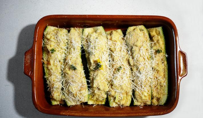 Fotografia em tom claro, com seis abobrinhas em fileira, recheadas e salpicadas com queijo ralado dentro de um refratário retangular marrom escuro, sobre a mesa branca.