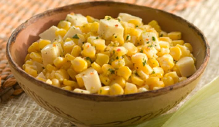 Fotografia em tons de amarelo, bege e rosa, com recipiente em tons de bege contendo milho verde refogado com salsa e pedaços de queijo, ao entorno espigas sobre base de palha marrom , palha de milho, tudo sobre toalha branca
